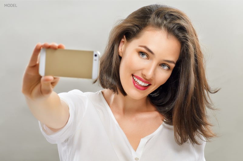 Woman taking selfie with gold cell phone against neutral background.