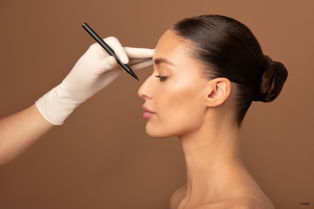 Young lady getting prepared for plastic surgery procedures by doctor drawing contours on her nose, standing on beige background, side view